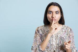 Confidential woman asking to keep a secret and be quiet in studio, putting index finger over lips. Indian girl being private and asking for silence, stand against blue background with mute sign. photo