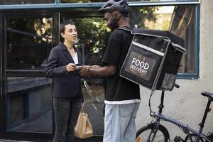 Woman client paying for her takeaway order with a contactless card at the doorstep. Delivery man carrying a thermal bag, completes fast food transaction using a wireless POS terminal. photo