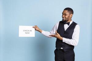 Professional bellboy showing a paper sign for guests to go to room check in, presenting an indicator to get the attention of clients. Hotel concierge in a tuxedo standing against blue background. photo