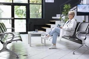 Older physician in hospital lobby examining x ray scans while sitting alone during appointment gap. Doctor in uniform reviewing patient materials in clinic hallway as part of evaluation process. photo