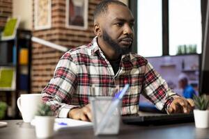 Focused black man working from home, reviewing company records on desktop pc. African american entrepreneur with hands on keyboard and mouse, updating business fiscal reports before deadline. photo
