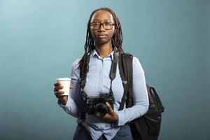 Young woman in suspenders and blue shirts, holding her camera with professionalism and creativity in photography studio. Black female photographer standing with coffee cup in hand. photo