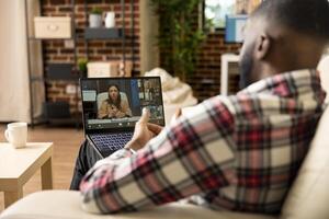 Black male entrepreneur presenting startup project plan to group of shareholders during virtual meeting. Businessman sitting on sofa with laptop, participating in online skills acquisition program. photo