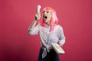 Furious young woman shouting into landline receiver. Angry caucasian lady yells at corded telephone, expressing frustration during bad call connection, isolated against pink background. photo