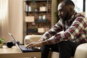Focused businessman working on laptop, preparing financial reports and meeting deadlines from cozy brick wall living room. African american male entrepreneur maintaining productivity in home office. photo