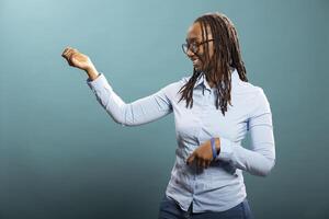 Smiling black woman with glasses having a good time in blue background studio. African american female individual standing and playing an imaginary guitar, feeling happy and optimistic for new day. photo