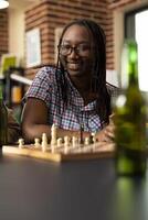 Selective focus on african american woman watching an ongoing friendly chess match on table. Smiling black lady with glasses sitting and enjoying board game session in brick wall room. photo