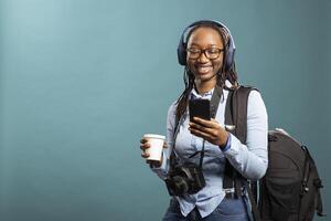 Joyful black woman with headphones watching funny videos on smartphone as she stands with coffee cup in hand and dslr camera around her neck. Female photographer using mobile phone with a smile. photo