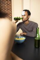 Young black man sipping his drink during fun indoor hangout with friends. African american guy seated at table with playing card on forehead, drinking beer and enjoying bonding moments with buddies. photo
