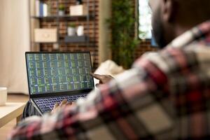 Remote trader sitting in living room, tracking stock indexes and cryptocurrency data on laptop. African american man working from home, reviewing trading market trends and investment strategies. photo