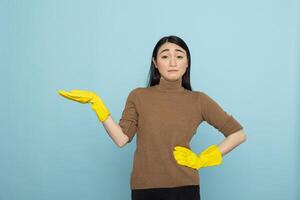 Asian housekeeper in yellow rubber gloves posing with one hand on waist and another gesturing to left side. Female cleaner presenting isolated copy space on blue background for promotional content. photo