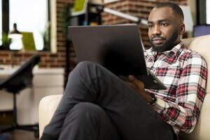 African american businessman seated with legs crossed, working from home, preparing startup project reports. Focused freelancer uses laptop on couch, updating resume and browsing job opportunities. photo