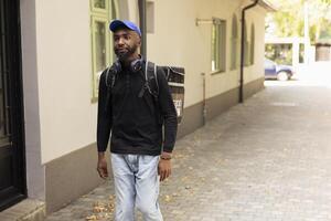 Black male courier approaches the front door to drop off a hot meal order, ringing the intercom to deliver the food. Young adult employee ensuring an express delivery takeaway service. photo