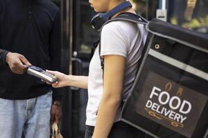 Black male client paying delivery bill with a card at pos during a takeaway drop off. Courier with backpack confirms the electronic payment for the completed meal delivery service. photo