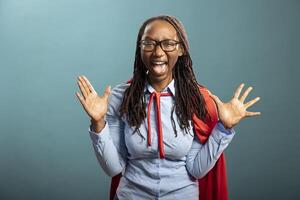 Confident black woman wearing a red cape and blue shirt, expressing authentic excitement in modern studio. African american heroine with her hands and mouth wide open, shouting with joy. photo