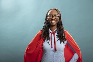 African american woman with red cape posing in blue background studio and confidently smiling at camera. Portrait of black female superhero standing strong, feeling empowered. photo