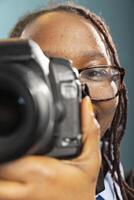Smiling african american woman holds DSLR close to her face, preparing to take a shot. Closeup of black female photographer showing how to use digital camera, set against a blue background. photo