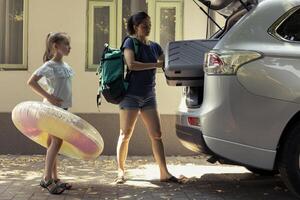 Mother and daughter smile while loading suitcase into the car for a weekend road trip. With the packed trunk, their holiday begins with laughter and adventure, excitement for the seaside. photo