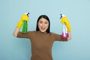Energetic asian housekeeper in rubber gloves holding colorful cleaning sprays and flexing arms, promoting cleanliness with big smile. Enthusiastic female cleaner with pink and green liquid solutions. photo