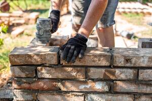 A worker wearing a glove and using a rubber mallet places a brick in place in a newly erected brick wall of a barbecue. photo