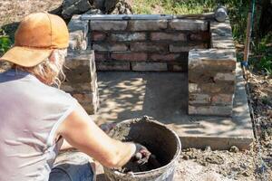 Man in cap mixing cement in bucket while building brick structure of outdoor barbecue photo