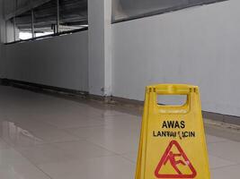 Yellow safety sign in Indonesian AWAS LANTAI LICIN, Caution Wet Floor, with a red falling figure symbol, indicating a slip hazard on a wet tiled floor in Banjarsari, West Java, Indonesia. photo