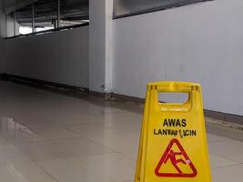 Bright yellow safety sign displaying AWAS LANTAI LICIN and a falling person icon, positioned on a reflective tiled floor, alerting to a wet and hazardous surface in Banjarsari, West Java. photo