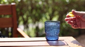 A hand squeezes lemon juice into a blue glass on a sunlit wooden table, creating a refreshing scene video