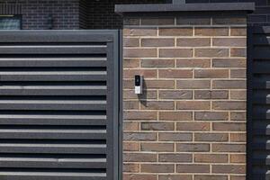A contemporary intercom is mounted on a brick wall adjacent to a slatted gate in a well-maintained suburban area photo