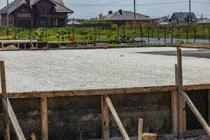 A concrete foundation has been freshly poured at a construction site surrounded by houses and greenery under a bright sky photo