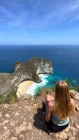 Woman enjoying breathtaking view of Kelingking Beach and turquoise ocean from cliff viewpoint on Nusa Penida, Bali. Perfect for travel, freedom, summer, and adventure themes. video
