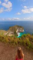 Cinematic FPV drone shot of woman in red dress walking toward dramatic cliffs and turquoise waters at famous Kelingking Beach on Nusa Penida island, Bali, Indonesia video