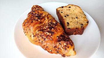 Delicious close-up of a croissant with chopped nuts and a slice of chocolate chip banana bread on a white plate. Perfect for bakery promotions, breakfast menu designs, or food blog visuals.. photo