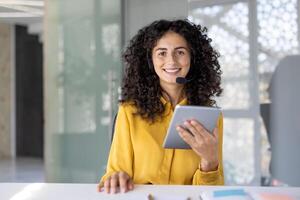 A smiling woman with curly hair wearing a headset, holding a tablet, looks directly at the camera in an office setting. photo