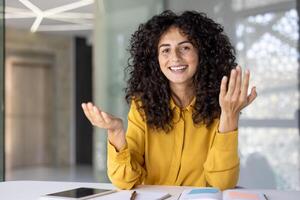 A cheerful woman with curly hair, gestures expressively while smiling, possibly presenting or explaining something in an office setting. photo