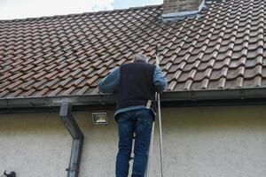 Professional roofer cleaning residential gutter while balancing on metal ladder, wearing work attire and checking roof condition under bright sunlight photo