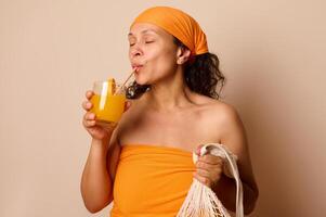 Woman Enjoys Fresh Orange Juice While Holding a Reusable Net Bag Against a Neutral Background photo