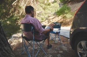Man Relaxing Outdoors in a Camping Chair by a Forest Road on a Sunny Day photo