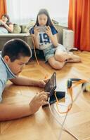 boy lying on wooden floor playing with tablet while two girls relax on sofa using smartphones with orange curtains and daylight, technology education leisure, vertical photo