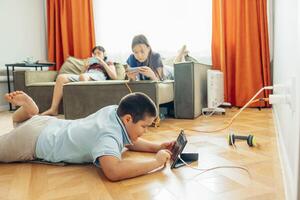 boy lying on wooden floor playing with tablet while two girls relax on sofa using smartphones with orange curtains, technology education leisure, power outlet with charging cables photo