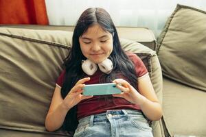 smiling Asian girl lying on green sofa using smartphone with headphones around neck focused expression home interior, light from window technology gadgets, lifestyle photo