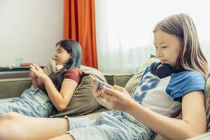 two girls relaxing on sofa using smartphone, headphones, casual home setting bright curtains and natural light window modern lifestyle, technology education entertainment photo