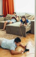 blurred boy lying on wooden floor playing with tablet while two girls relax on sofa using smartphones with orange curtains and daylight, technology education leisure, vertical photo