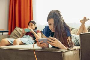 Asian half-breed girl on sofa smiling, playing on smartphone, headphones, orange charging cable connected, blurred background, sister, friend, technology education entertainment photo