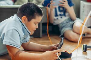 boy lying on wooden floor playing with tablet while Unrecognizable girl relax near sofa using smartphones with orange curtains and daylight, technology education, game photo
