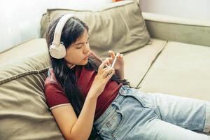 Asian teen girl lying on green sofa using smartphone with headphones, focused expression home interior, light from window technology gadgets, lifestyle photo