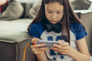 close up of Asian half-breed girl near green sofa using smartphone, headphones, home interior with soft light from window technology gadgets youth lifestyle photo