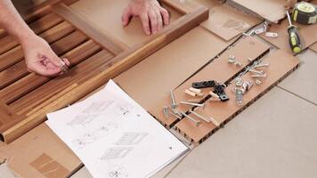Hands assembling wooden furniture using tools and instructions. Bright lighting highlights detailed steps on a tiled floor video