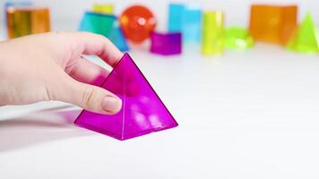 A hand manipulates a vibrant purple pyramid among various colorful geometric shapes on a white surface video