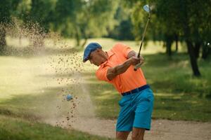 Golf player makes a powerful swing from a sand trap on a sunny day at a lush green golf course surrounded by trees photo